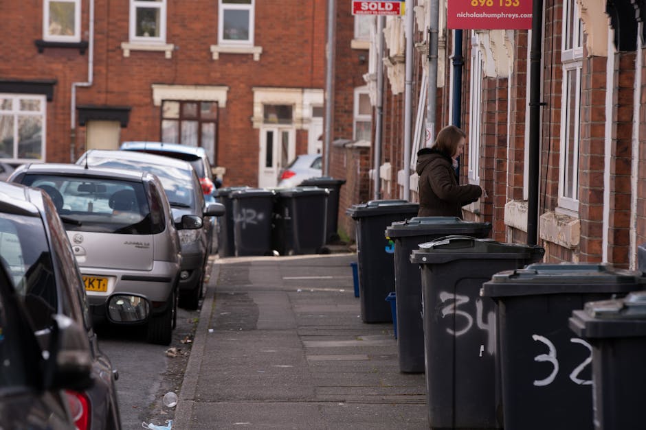 A row of dark grey wheelie bins with lids, lined up along a narrow urban residential sidewalk, each bin marked with white spray-painted numbers. The bins are positioned against the front brick facade