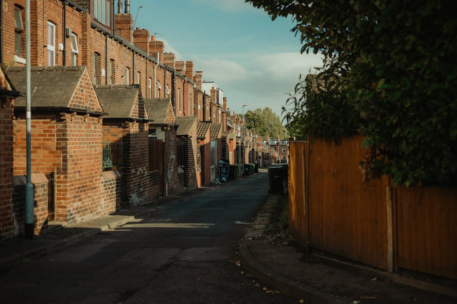 A row of dark grey wheelie bins with lids, lined up along a narrow urban residential sidewalk, each bin marked with white spray-painted numbers. The bins are positioned against the front brick facade